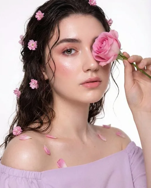 A hyper-realistic close-up portrait of a woman with a dreamy expression, holding a pink rose near her face, featuring wavy dark hair adorned with soft pink flowers and petals, set against a pure white background with soft diffused lighting.