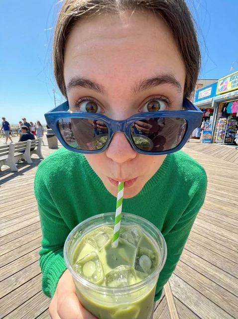 An ultra-wide fisheye close-up of a young person with a curious expression, wearing a bright green sweater and blue sunglasses, sipping an iced matcha latte on a sunny boardwalk with wooden planks and colorful stalls in the background.