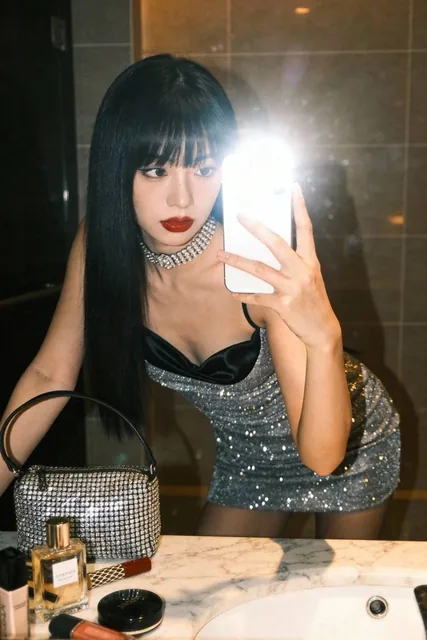 A stylish young Asian woman in a sparkly silver mini dress takes a mirror selfie with flash in an upscale hotel bathroom, featuring porcelain skin, red lips, and a chic expression, with makeup products and warm ambient light in the background.