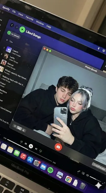 A hyper-realistic downward-angled shot of a MacBook screen with subtle imperfections like dust and reflections, displaying a Photo Booth window showing a couple in a dark bedroom wearing black hoodies, with cool screen glow and warm skin tones.