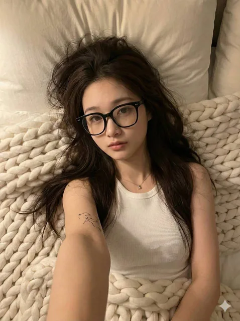 A young adult female with long dark brown tousled hair and light skin tone, wearing a white ribbed tank top and thick black-rimmed glasses, lying on a bed with a chunky knit white blanket and soft pillows, captured in a high-angle selfie with warm lighting and a relaxed mood.