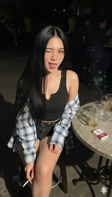 A young woman with fair skin, natural reddish tones, and freckles sits on a bar chair, winking and sticking out her tongue, wearing a black tank top, denim skirt, and plaid flannel shirt, holding a lit cigarette in a dark bar setting with harsh flash lighting.