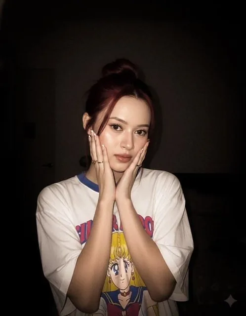A close-up photo of a young, beautiful, fair-skinned woman wearing a white oversized t-shirt with a large Sailor Moon image, in a dimly lit bedroom at night with a bright flash highlighting her face and short black hair, posing with a gun gesture and a cute expression.