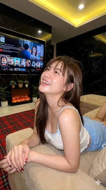 A young adult female with long brown hair and glossy lips, wearing a spaghetti-strap top in white and light blue, lying naturally on a plush sofa in an indoor living room with a blurred background featuring a TV showing Netflix logo, a modern fireplace, and dark marble walls, captured in a soft-focu