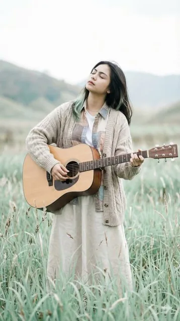 An ethereal full-body portrait of a beautiful young Asian woman standing in a vast meadow, strumming a light-wood acoustic guitar with intense emotion. She wears a chunky cable-knit cardigan and linen skirt, surrounded by tall wispy grasses under a pale overcast sky with soft blurred hills in the ba