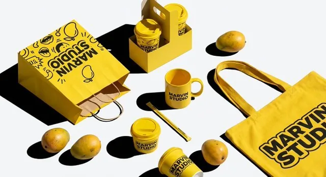 A high-contrast flat lay of Marvin Studio's Mango Fruit Tea brand identity collection, featuring vibrant yellow items like a shopping bag, ceramic mug, and fresh mangoes arranged in dynamic knolling on a white background with sharp shadows and distinct textures.