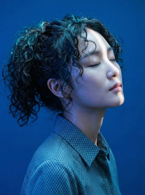 An ultra-realistic editorial close-profile portrait of a Korean woman with closed eyes and head tilted upward, immersed in dramatic cobalt blue lighting. A diagonal cold white beam precisely illuminates her facial features against a solid Prussian blue background, highlighting her monolid eyes, high