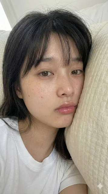 An extreme close-up portrait of an adult East Asian woman lying on her side with her cheek resting on a cream pillow, featuring natural skin texture with light freckles, glossy pink lips, and direct eye contact in soft, diffuse lighting.