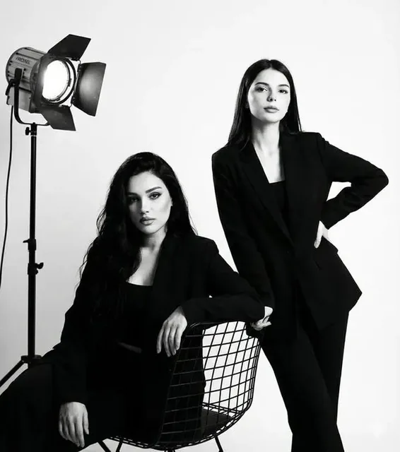 A high-contrast black and white studio portrait of two young Asian women. One is seated on a minimalist wire chair in the foreground, wearing a black blazer and looking intensely at the camera. The other stands behind, leaning in gently with a composed expression. Dramatic cinematic lighting creates
