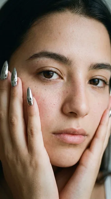 An extreme close-up editorial macro portrait of a woman in her early 20s with natural skin texture, freckles, and minimal makeup, resting her hands against her face. The image features real window lighting, subtle film grain, and a focus on authentic human details.
