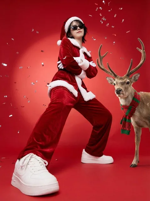 A person in a rich red velvet Santa suit with white fur trim and sunglasses strikes a confident swagger pose in a bright red studio. A realistic reindeer with a knitted scarf stands beside them as silver confetti floats in the crisp commercial lighting.