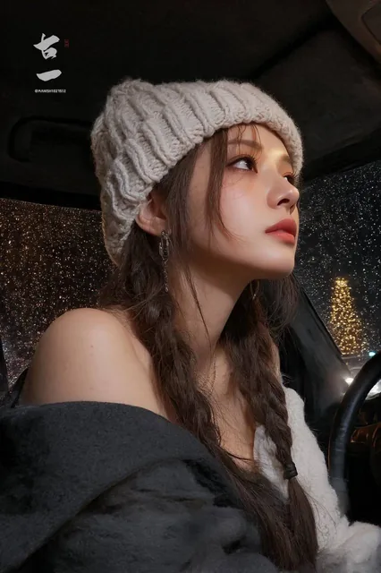 A young East Asian woman in her early 20s sits in the driver's seat of a car on a rainy night, her head slightly lifted as she looks out the window with a calm, introspective expression. She wears a cream cable-knit beanie with long braids, a black coat slipping off one shoulder, and delicate silver