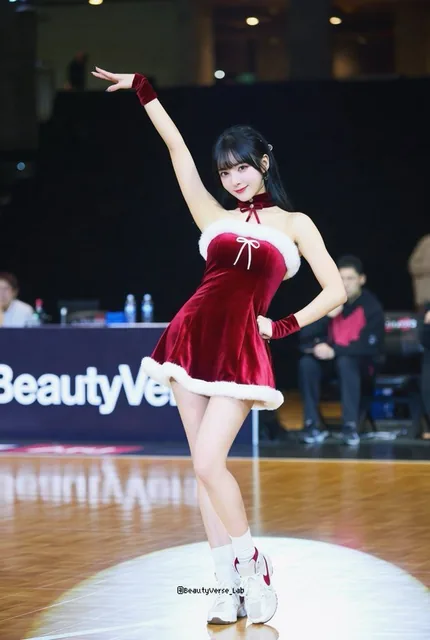 A confident K-pop idol posing courtside in a festive red velvet Santa mini dress with white fur trim, standing on glossy hardwood flooring with strong reflections, against a softly defocused background featuring 'BeautyVerse' signage.