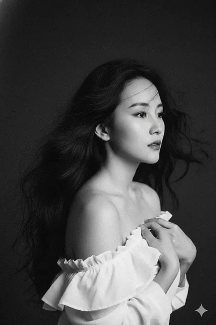 A black and white artistic portrait of a beautiful woman with classic features and wind-blown wavy black hair, holding white fabric against her chest with an off-shoulder pose, against a dark background with pronounced film grain texture.