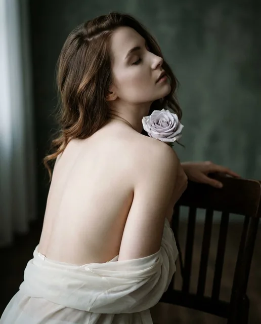 A young woman with auburn hair in a 3/4 rear view, wearing a sheer ivory robe off her shoulders, holding a pale lavender-gray rose near her collarbone. She leans forward with eyes closed in a moody indoor setting with soft side lighting and a dark green-gray mottled wall.