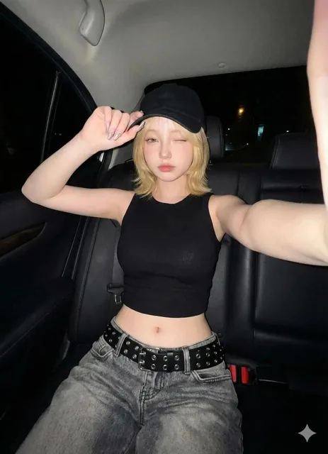 A young woman with a butterfly haircut takes a selfie in the back seat of a car at night using an iPhone 17 Pro. She wears a black ribbed sleeveless crop top and low-rise faded gray baggy jeans with a wide black belt. Her right hand holds the phone while her left hand touches the tip of her black ca