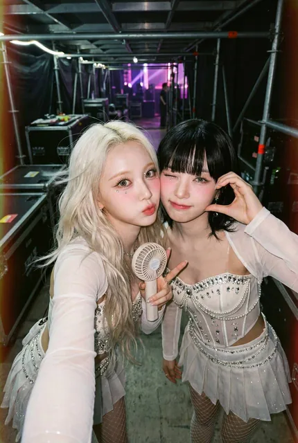 Two stunning K-pop female idols in high-concept stage outfits pose cheek-to-cheek for an intimate backstage selfie. One has platinum blonde waves, the other a jet black hime-cut. They are in a backstage corridor with blurred equipment, captured with a soft-focus analog film camera aesthetic, heavy g