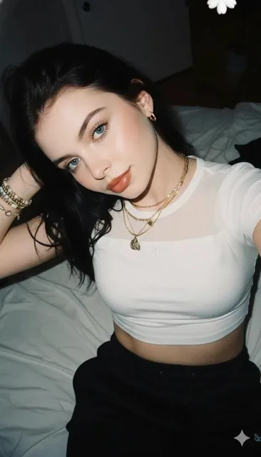 A playful yet confident selfie portrait with direct flash lighting, featuring a woman with long black messy hair lying sideways on a bed, wearing a white crop top and black sweatpants, in a casual 2000s aesthetic style with warm skin tones and a flat background.