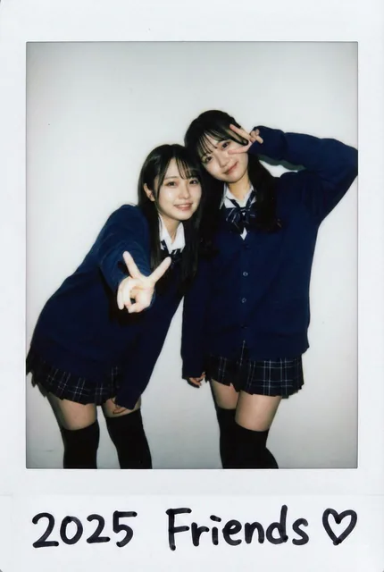 Two young Japanese women in matching JK school uniforms pose closely for a portrait. One leans forward making a peace sign, while the other tilts her head affectionately. The image has a harsh on-camera flash, a plain background, and a thick white border with handwritten text '2025 Friends ♡', mimic