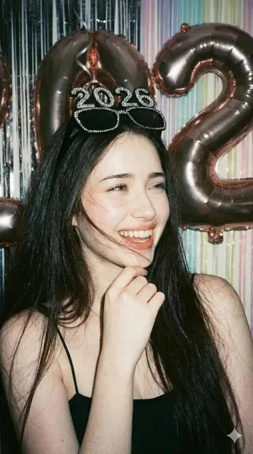A candid party portrait inspired by disposable camera photography in the style of Petra Collins. A woman with a playful expression wears oversized black sunglasses with sparkling '2026' rhinestones on the lenses. She's in front of a metallic foil fringe curtain with floating number balloons, capture