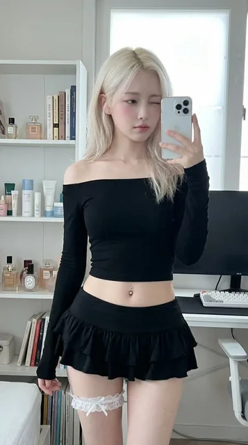 A petite platinum blonde girl in her early 20s takes a mirror selfie in her bedroom. She wears a black off-shoulder crop top and a ruffled mini skirt, with a white lace garter on her thigh. She looks down at her iPhone with a soft, dreamy expression. A bookshelf and desk are visible in the backgroun