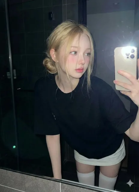 A young woman stands in a slightly dark bathroom, leaning forward slightly and looking at the mirror. She wears an oversized black t-shirt, a short white skirt, and thigh-high socks. The photo is taken with direct flash, capturing soft makeup, curled eyelashes, and natural hair with some strands on