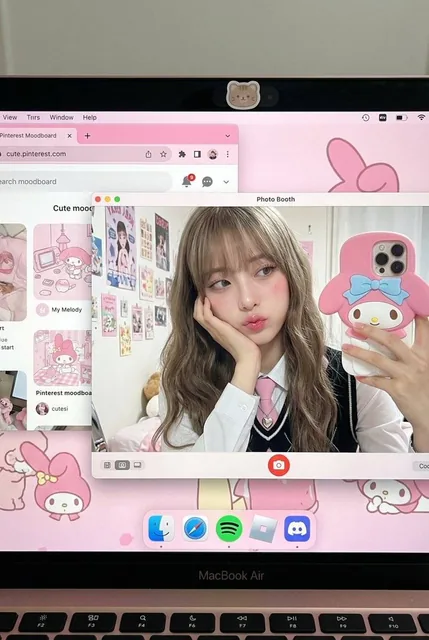 A photorealistic close-up POV shot of a powered-on MacBook Air screen. The pink desktop shows a Pinterest moodboard and a Photo Booth window with a girl taking a selfie. She has long wavy hair, wears a school uniform, and holds a My Melody phone case. The image uses direct flash for a hard light eff