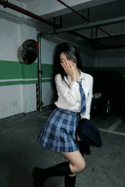 A young woman in a blue plaid school uniform shyly covers her face with her hand while standing in a dimly lit underground parking garage, captured with motion blur and a candid smartphone aesthetic.