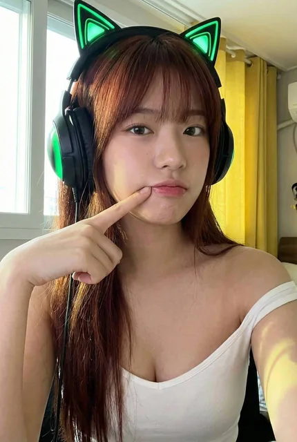 A photorealistic selfie portrait of a girl wearing a green LED cat ear gaming headset, pulling her lower lip with her finger, illuminated by natural window light from the left with a yellow curtain background.