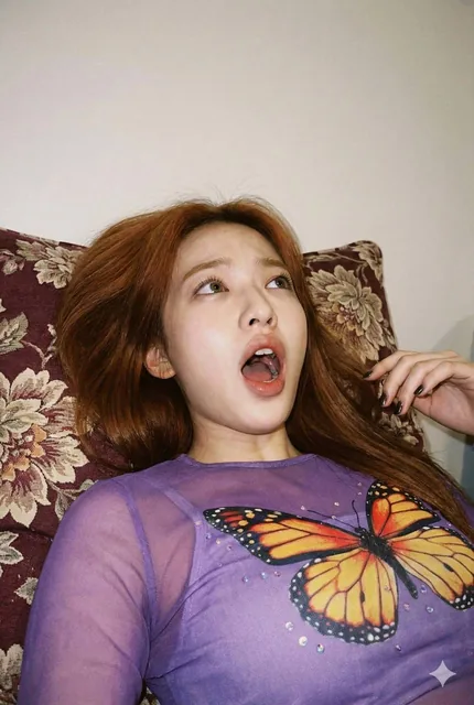 A close-up, low-angle portrait of a young blonde woman with a dramatically shocked expression, mouth agasp and eyes rolled upward. She wears a purple sheer top with a colorful butterfly print and lies against a patterned pillow. The image has a soft, slightly grainy, candid smartphone photography ae
