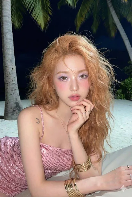 A female with doe eyes and strawberry blonde hair reclines on a tropical beach at night, wearing a pink sequined tube dress. She has a soft-glam makeup look with lilac eyeshadow and rosy blush, and a small crescent moon tattoo on her shoulder. The scene is lit by a soft frontal flash and cool ambien