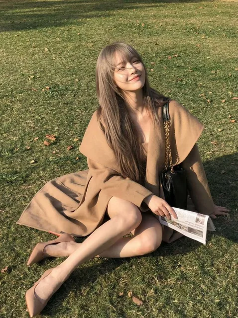 A woman in a camel coat enjoys a lazy afternoon, with wind tousling her hair and sunlight bathing her. Shot from a low angle with a 24mm wide-angle lens to elongate her legs, creating a relaxed and cinematic vibe with Fujifilm NC filter tones.