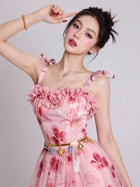 A high-fashion editorial portrait of a confident female model with smooth pinkish-white skin and gray-blue contact lenses. She wears a flowing abstract floral chiffon dress in pink and red tones with a decorative petal-like neckline and bow-tie straps, accessorized with a gold waist belt featuring s