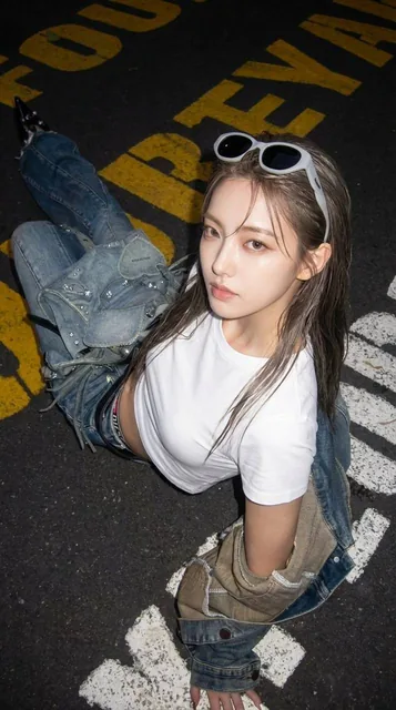 A dynamic ultra-wide-angle flash photograph taken from a high angle at night, featuring a young woman sitting on gritty asphalt. Her face, with detailed makeup, is large and prominent on the right side of the frame, while her body angles away towards the left, creating a dramatic forced perspective