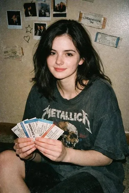 A young woman with pale skin and dark brown hair sits on the floor, wearing an oversized faded band T-shirt. She holds concert tickets and gives a subtle smile in a dim room with a beige wall covered in Polaroids, doodles, and tickets. The image has the raw, flat-shadowed look of a 90s disposable ca