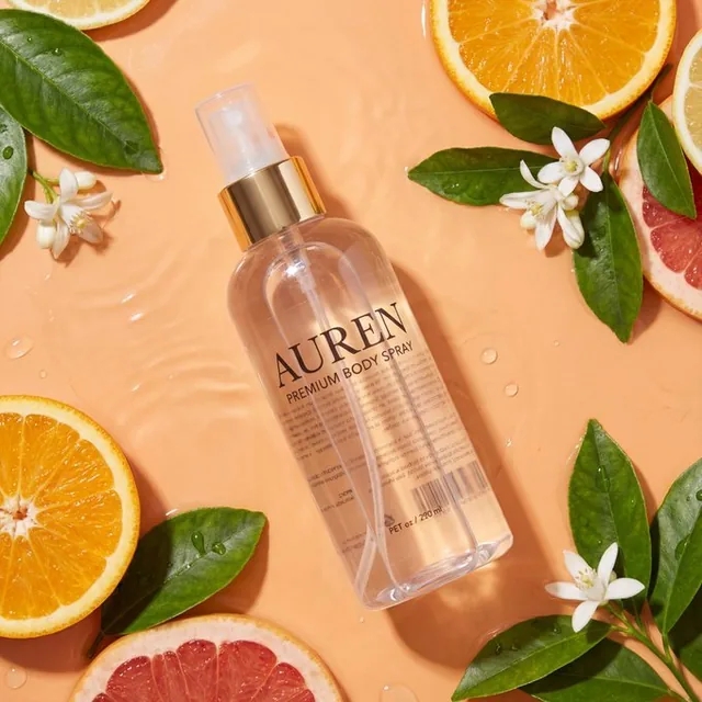 Ultra high-end photorealistic studio product shot of an AUREN body spray bottle surrounded by fresh botanical elements and citrus fruit on a peach-orange wet surface.