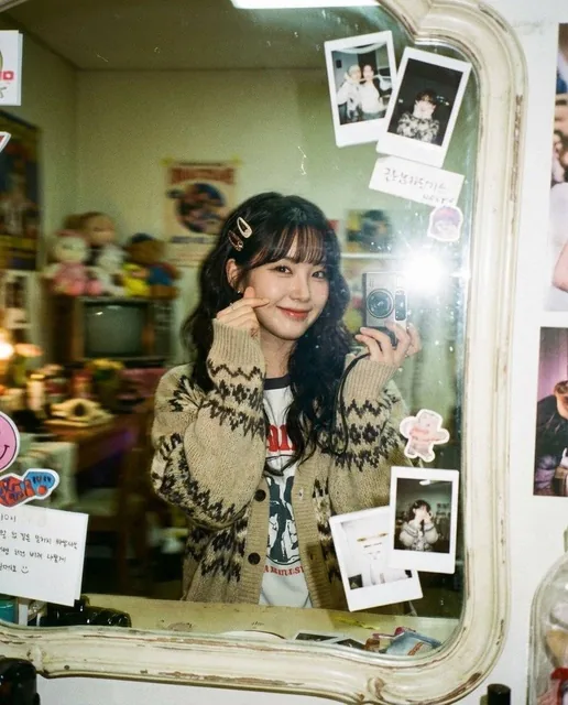 A young cute Korean K-pop idol making a finger heart gesture near her cheek, smiling warmly at her reflection in a vintage mirror covered with stickers and polaroids. She wears an oversized colorful knit cardigan in a cozy, cluttered room. The image has a 90s lo-fi film aesthetic with heavy motion b