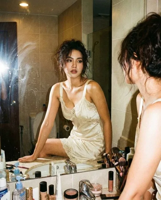 A raw candid flash-style mirror photograph of a young woman in a vintage silk slip dress, seated on a cluttered bathroom vanity. The image uses the exact face identity and visible text from the uploaded reference image, captured with harsh on-camera flash and warm ambient tungsten lighting.