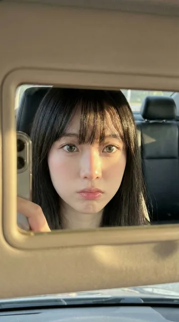 An ultra-photorealistic close-up portrait of a young East Asian woman with pale porcelain skin and deep black hair, reflected in a car sun visor vanity mirror during golden hour. The photo captures a candid, intimate moment with sharp side lighting creating strong highlights and abstract hair shadow