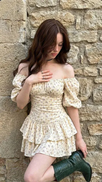 A young woman with long dark chestnut hair leaning against a textured stone wall, wearing a cream floral off-the-shoulder dress and holding a dark green crocodile-texture cowboy boot, captured in soft natural daylight with an artistic, moody aesthetic.