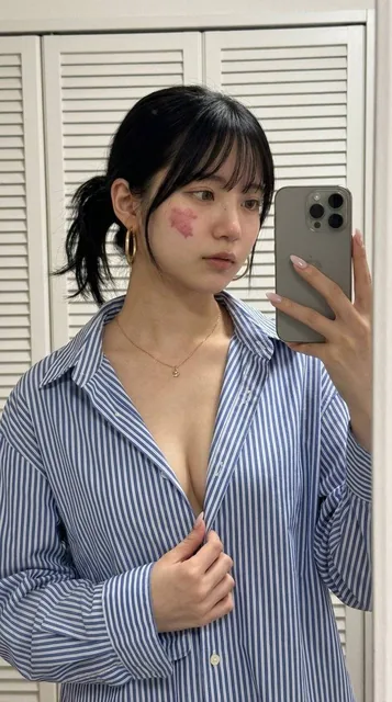 A realistic portrait of a platinum blonde woman in her mid-20s taking a mirror selfie. She wears an oversized blue-and-white striped button-down shirt, unbuttoned and held open with one hand. She has an athletic build, almond-shaped hazel-green eyes, and a calm, confident expression. The background