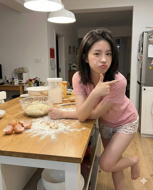 An ultra-realistic portrait of a young athletic woman with fair skin playfully pouting in a messy kitchen. She has a smudge of white flour on her nose and cheek, slightly messy short-to-medium hair, and is wearing a plain pink t-shirt and tiny pajama shorts. She leans over a kitchen island counter w