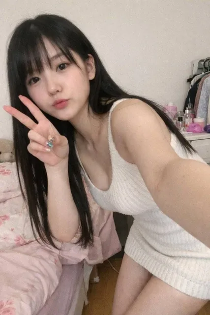 A young woman with long straight black hair takes a casual selfie in her delicate bedroom. She wears a tight white knitted dress and has a slim figure with cool-toned fair skin. The photo has an offhand, iPhone-like snapshot quality with random composition.