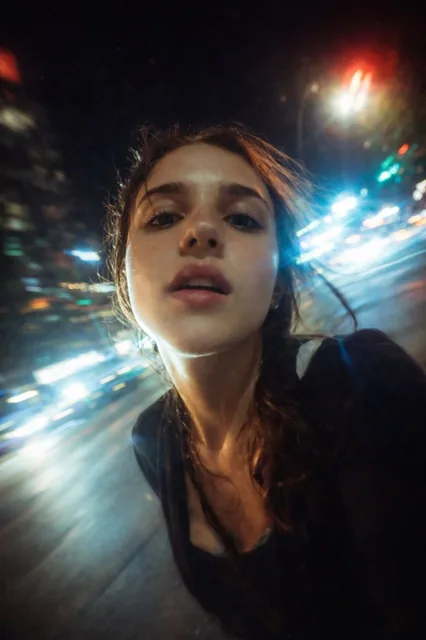 A cinematic night-time selfie of a young woman riding through a cyberpunk city, captured from a low wide-angle perspective with soft motion blur, neon light trails, and a grainy 35mm film texture.