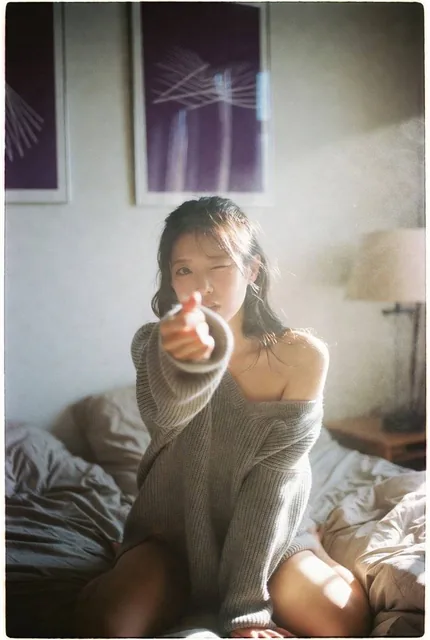 A young Korean woman with a K-pop idol aesthetic sits on a rumpled bed, facing the camera with a playful, sexy-cute expression. She wears an oversized knit sweater off one shoulder. Warm sunlight illuminates the scene, creating a hazy, overexposed look with heavy film grain. Framed abstract purple p