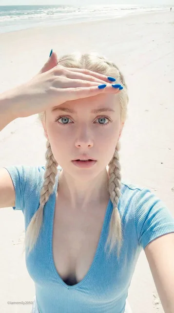 A young woman with porcelain doll features takes a high-angle selfie on a bright sandy beach. She shields her eyes with one hand against the coastal light, wearing a light blue deep V-neck top and platinum blonde braids, with a neutral, vacant expression.