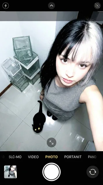 A raw, unedited smartphone selfie taken from a high-angle perspective looking straight down at a young woman with split-dyed hair and a black cat at her feet, in a bright room with glossy ceramic tiles and geometric acrylic cubes, featuring realistic lighting artifacts and camera distortions.