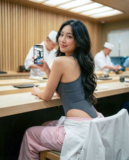 A candid lifestyle photo of a young Asian woman with long wavy dark hair looking back over her shoulder with a playful smile while seated at a high-end sushi restaurant. She wears a charcoal grey bodysuit and pastel pink track pants, holding a smartphone. The background features creamy bokeh with ve