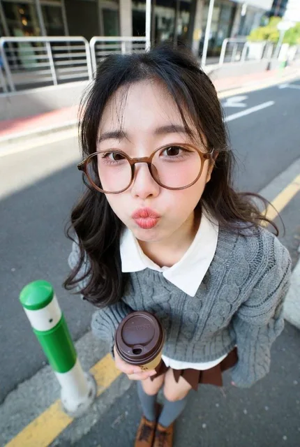 A young woman with dark brown wavy hair wearing round brown glasses and a gray cable-knit sweater over a white shirt, holding a takeaway coffee cup with both hands while leaning forward on a city sidewalk with modern buildings in the background.