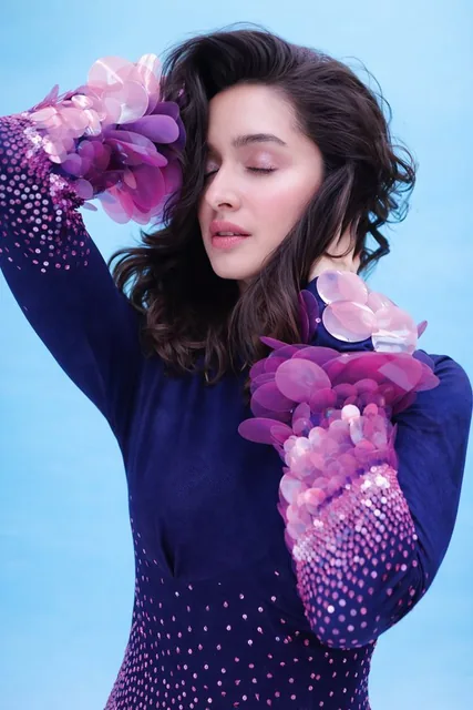 A young adult female stands in a studio, wearing a form-fitting, long-sleeve navy blue dress adorned with reflective pink, magenta, and purple circular appliqués and sequin-like el